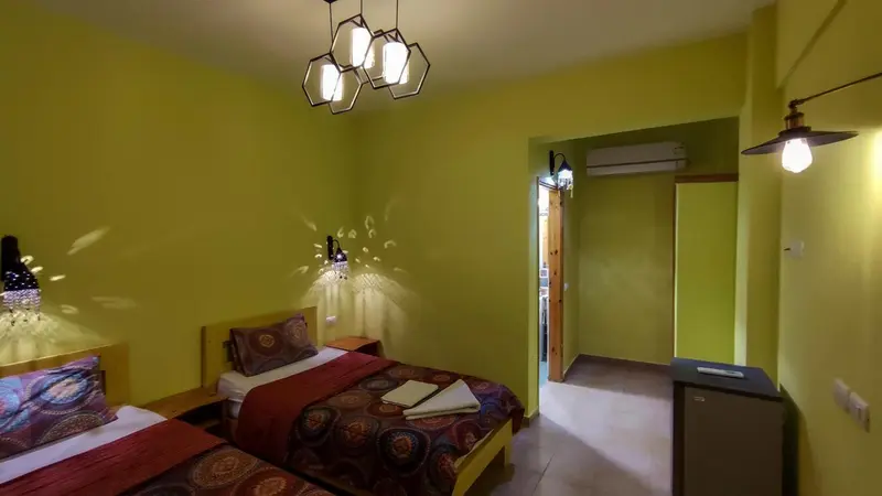 Une chambre aux murs verts et au lit recouvert d'une courtepointe colorée est éclairée par un lustre moderne.
