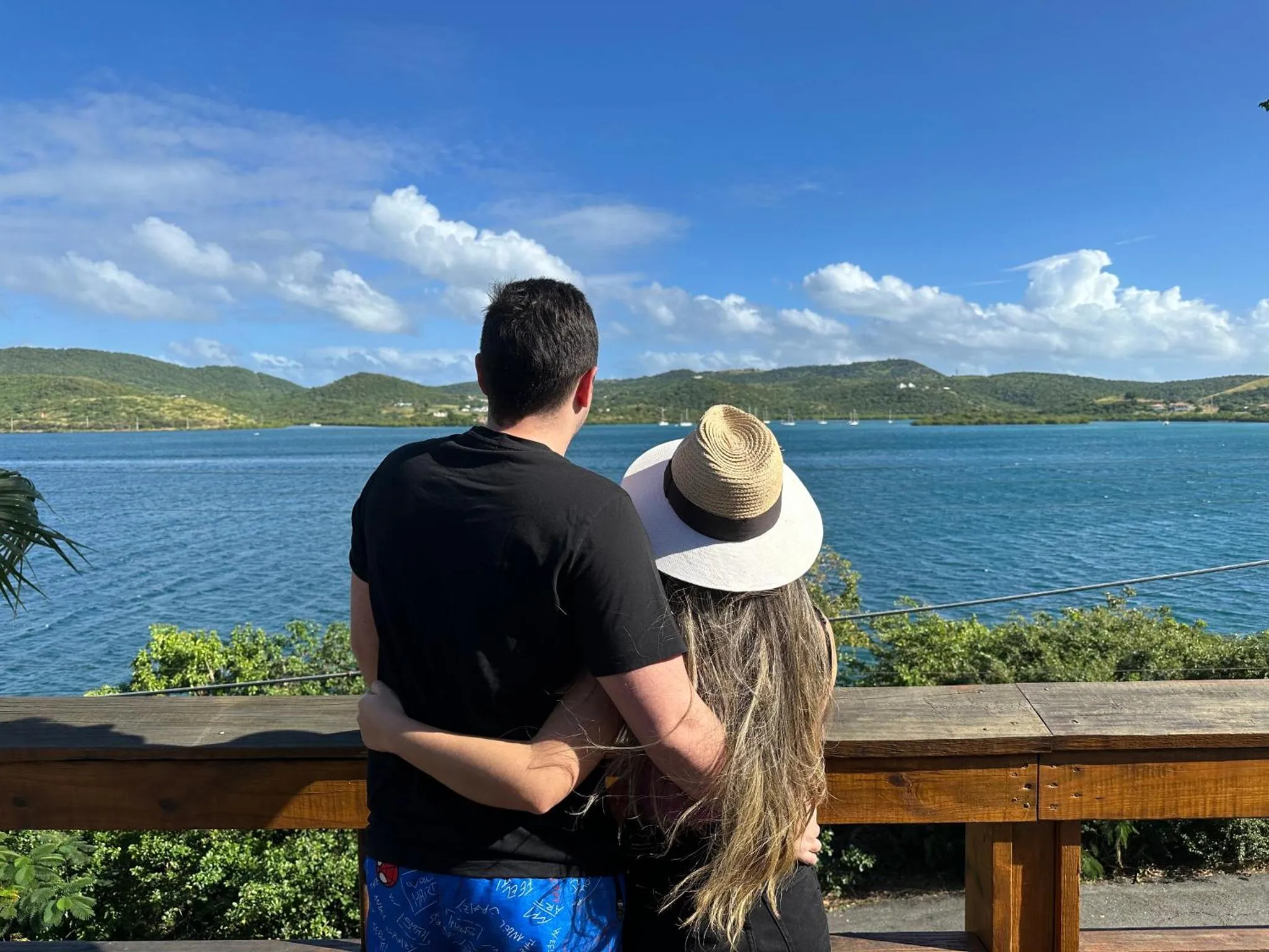 Un couple se tient dos à la caméra, contemplant une vue sur la mer et les collines.