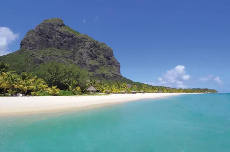 Plage de sable blanc bordée d'une eau turquoise et d'une végétation luxuriante au pied d'une montagne rocheuse.
