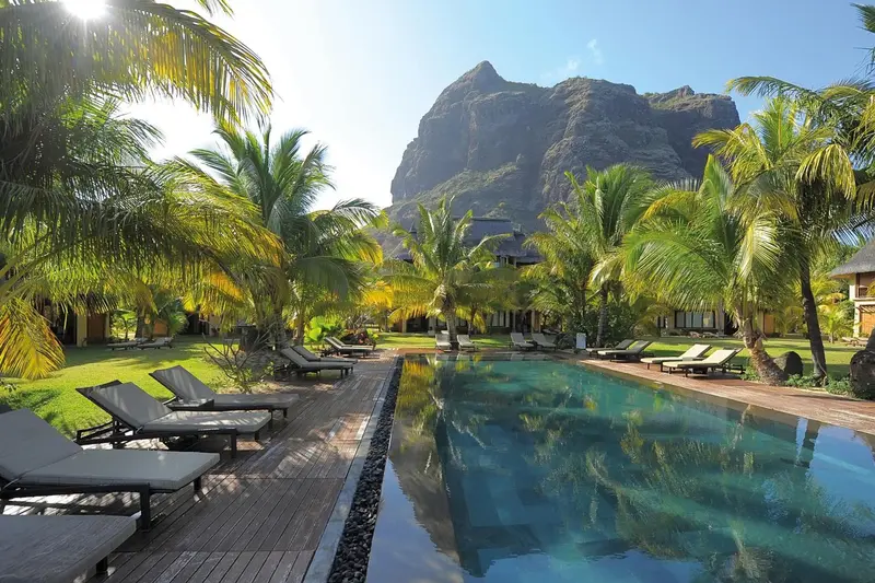 Transats alignés au bord d'une piscine turquoise avec une végétation luxuriante et une montagne en arrière-plan.