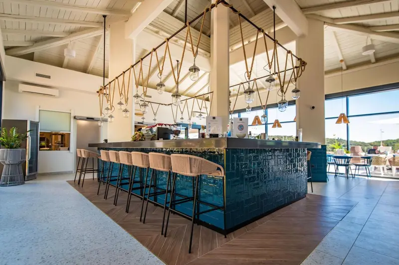 Un long comptoir de bar turquoise avec des tabourets en osier est éclairé par des suspensions décoratives.