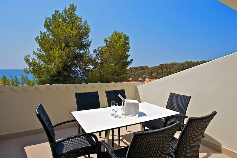 Une table et des chaises noires sont disposées sur une terrasse lumineuse avec vue sur la mer et la végétation.