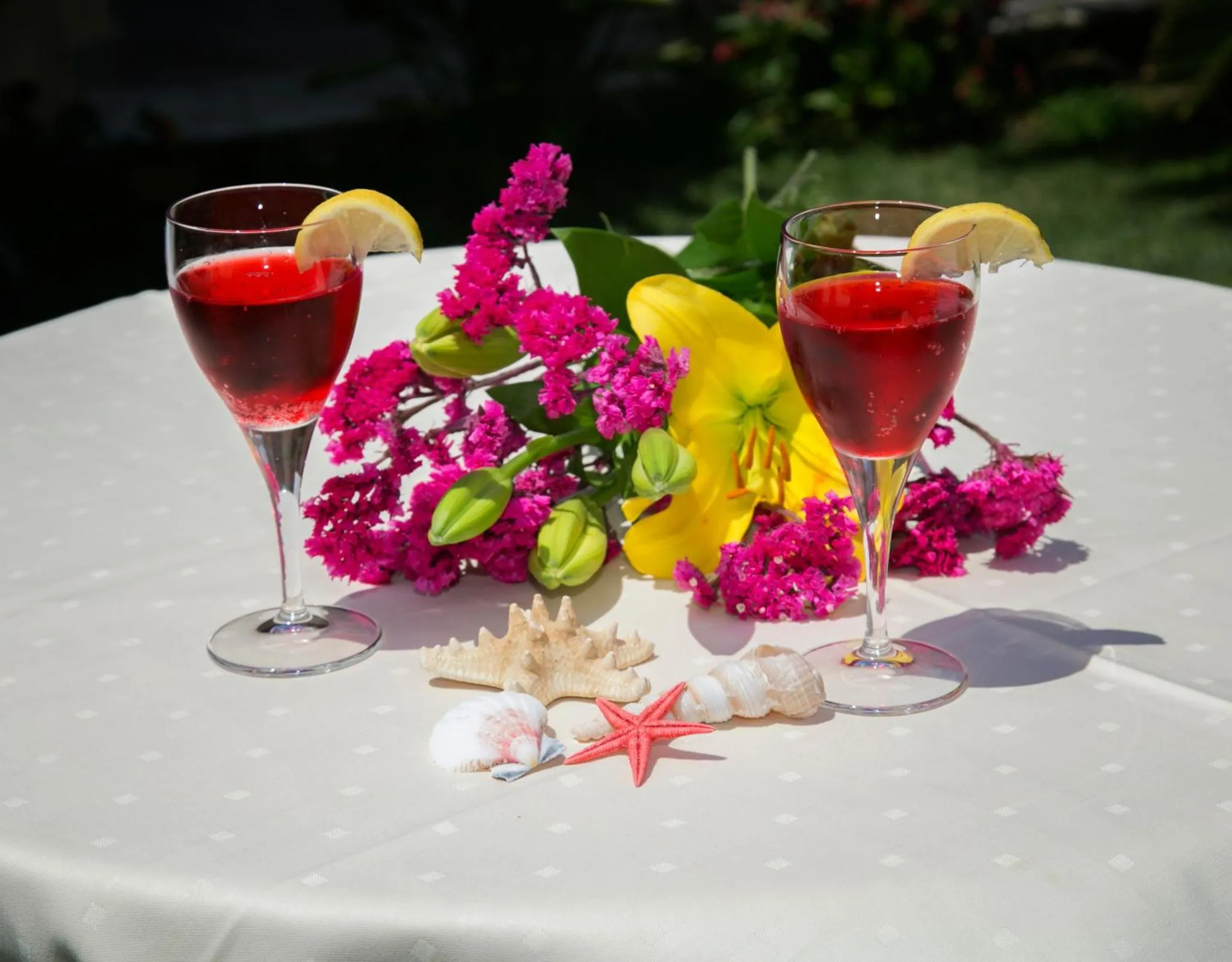 Deux verres de boisson rouge garnis de citron et entourés de fleurs et de coquillages sur une table.
