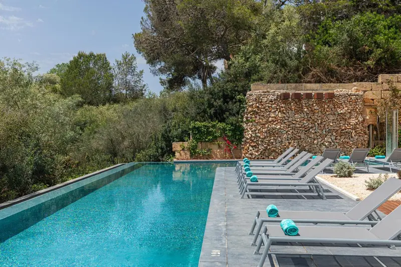 Une piscine rectangulaire bordée de chaises longues turquoise et d'une murette en pierre.