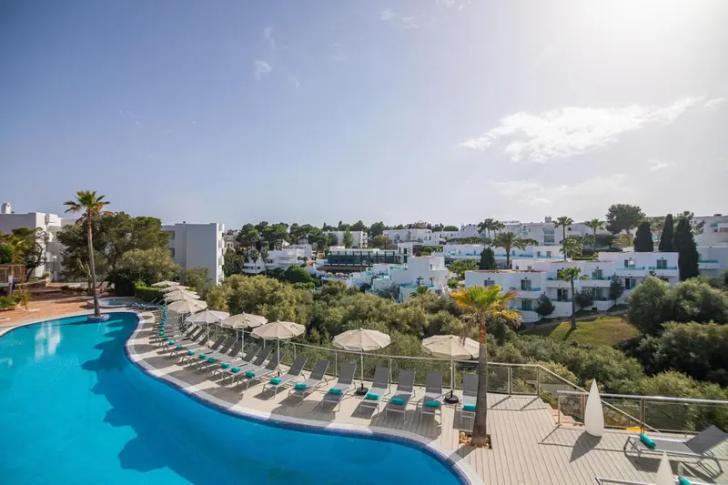 Une piscine bleue bordée de transats et de parasols offre une vue sur un ensemble de bâtiments blancs et de végétation luxuriante.