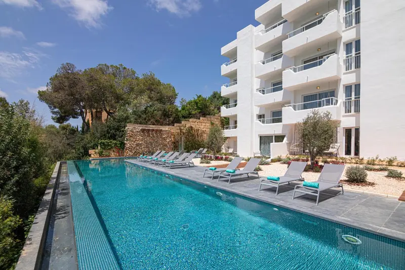 Une piscine rectangulaire bordée de transats se trouve devant un bâtiment blanc à balcons.
