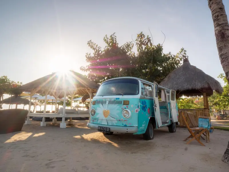 Un combi Volkswagen coloré se trouve sur une plage de sable fin, avec des parasols et des abris de plage en arrière-plan.