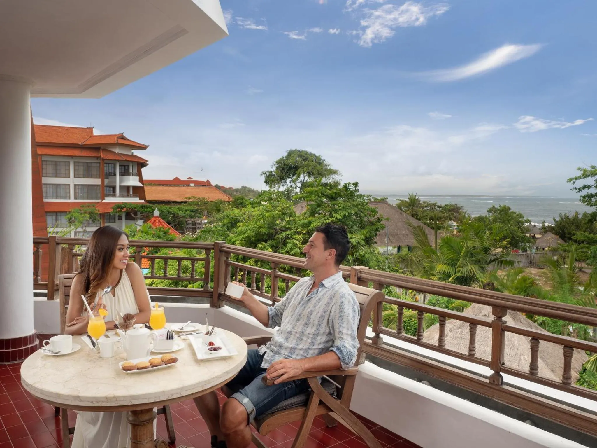 Un couple prend son petit-déjeuner sur une terrasse ensoleillée avec vue sur la mer et la végétation.