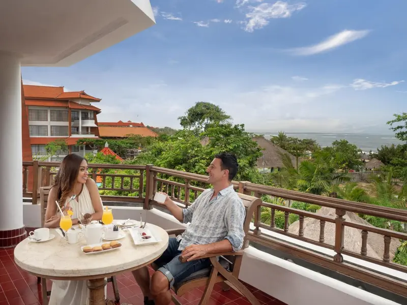 Un couple prend son petit-déjeuner sur une terrasse ensoleillée avec vue sur la mer et la végétation.