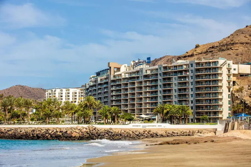 Bâtiment blanc avec de nombreux balcons surplombant une plage de sable et une mer bleue.