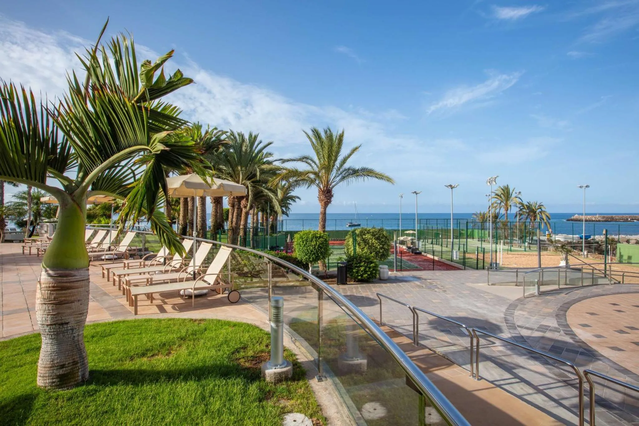Terrasse ensoleillée avec transats et palmiers, vue sur la mer et un court de tennis.