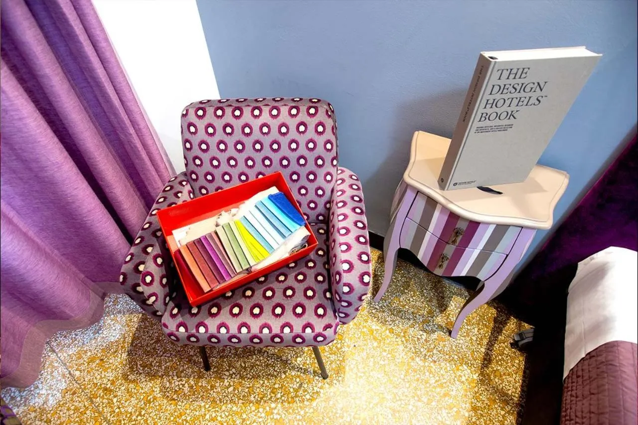 Un fauteuil violet à motifs se trouve près d'une table de chevet rose et d'une pile de livres.