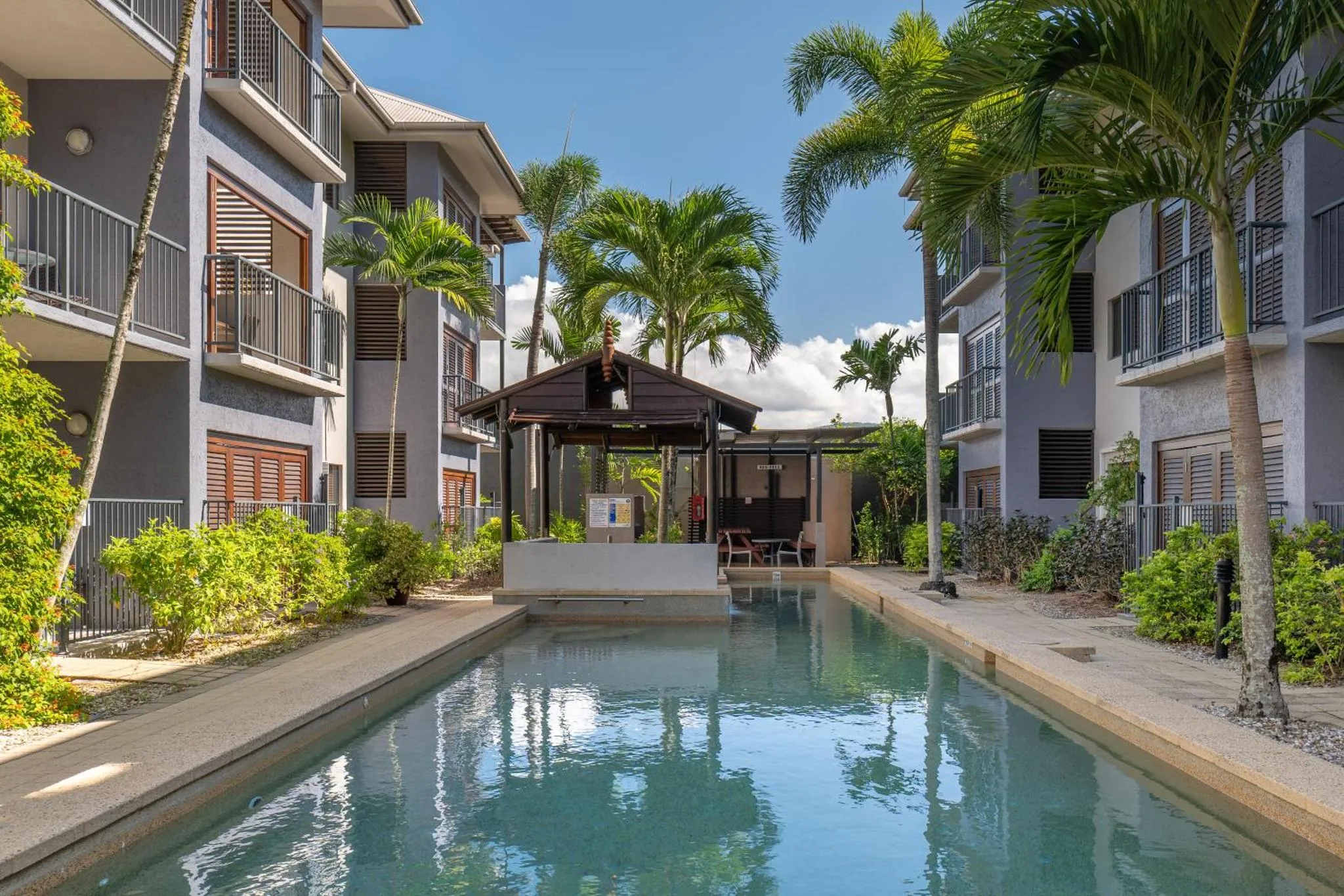 Une piscine rectangulaire bordée d'une terrasse ensoleillée, entourée de bâtiments résidentiels et de végétation tropicale.