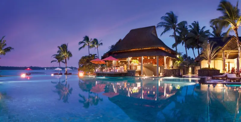 Une piscine à débordement reflète les lumières d'un bâtiment tropical au crépuscule.