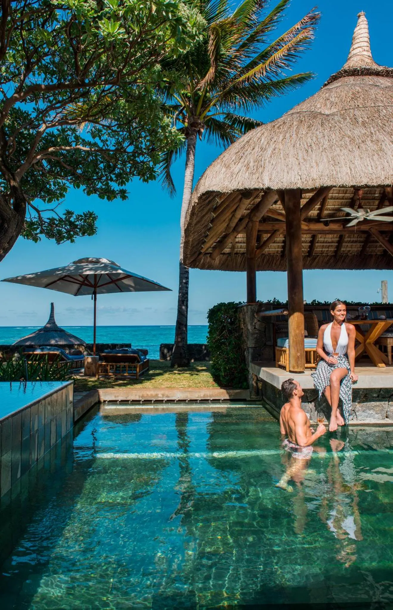 Un couple se détend près d'une piscine avec un abri de chaume et une vue sur la mer.