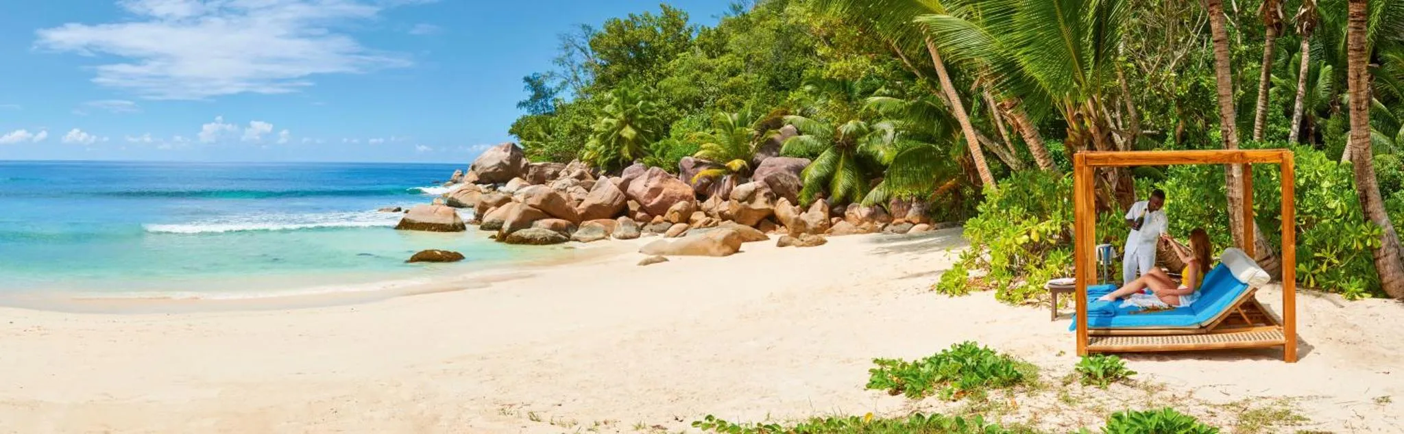 Deux personnes se détendent sur des transats sur une plage de sable blanc bordée de rochers et de végétation tropicale.
