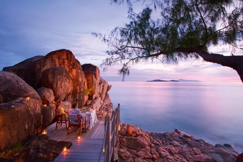 Une table est dressée pour un dîner en extérieur avec vue sur la mer au coucher du soleil.