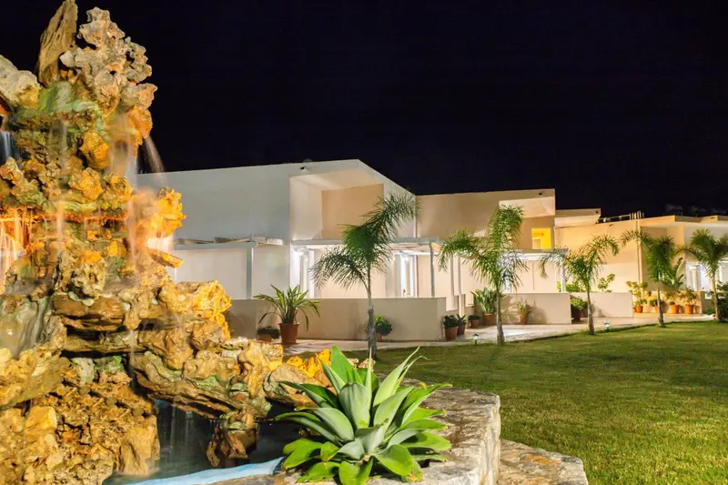 Une fontaine illuminée orne un jardin soigné devant un bâtiment clair.