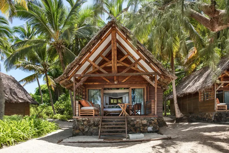 Petite maison en bois sur une plage de sable blanc bordée de palmiers.