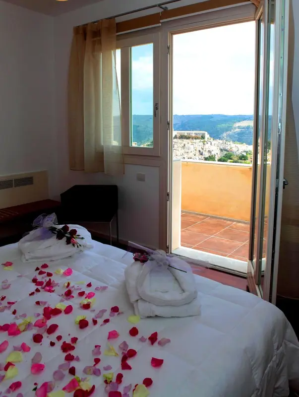Une chambre lumineuse avec un lit décoré de pétales de fleurs donnant sur une terrasse et un paysage montagneux.