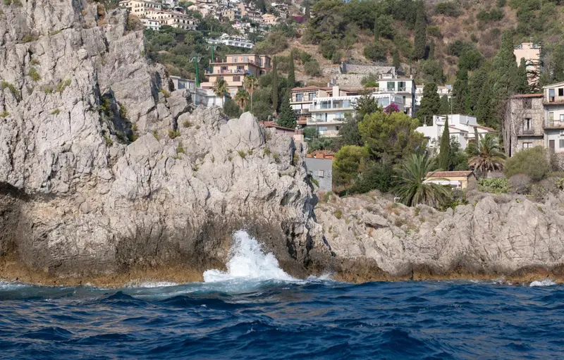 Vue côtière rocheuse avec des vagues s'écrasant et des bâtiments en arrière-plan.