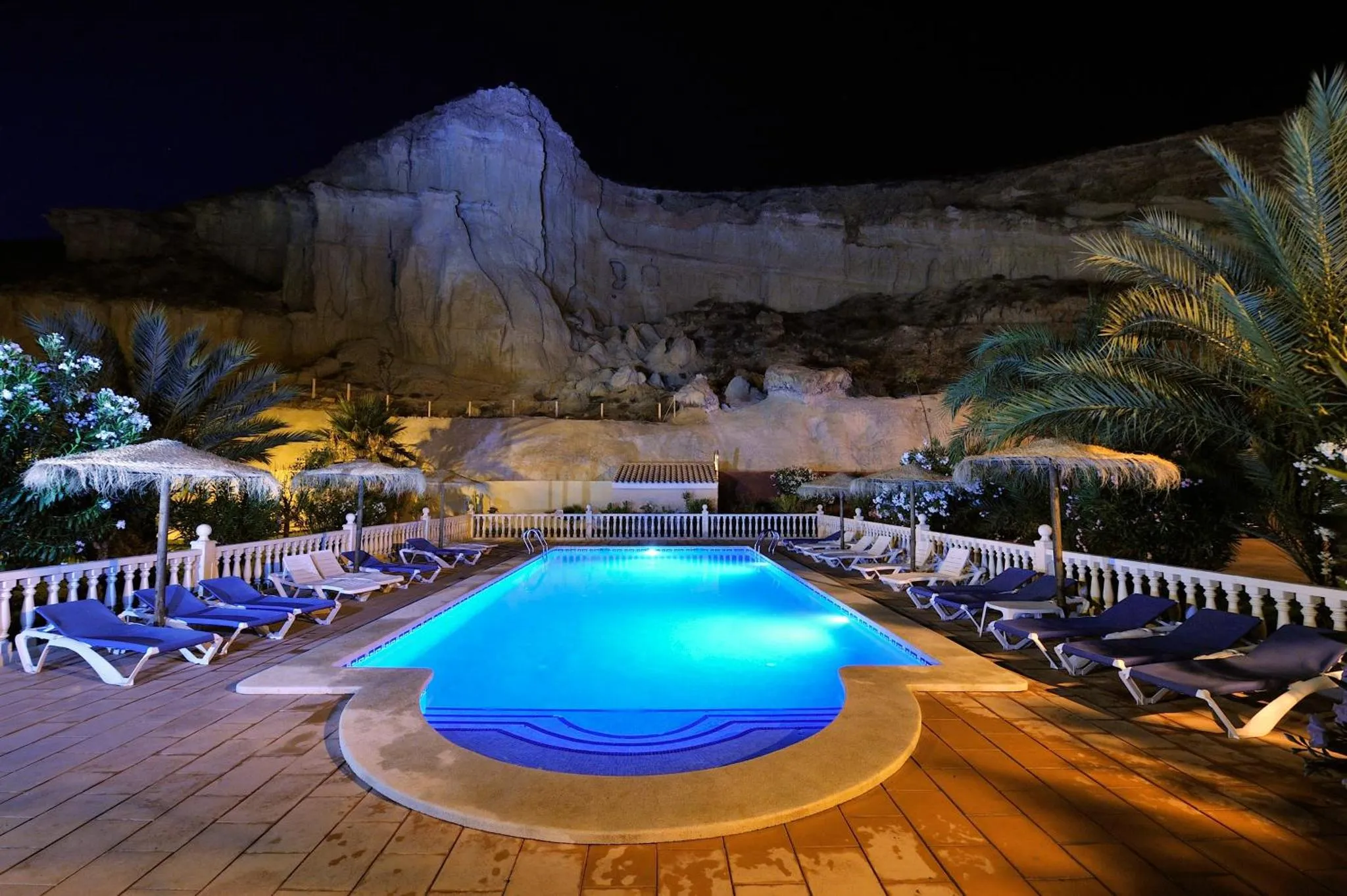 Piscine illuminée avec transats et végétation luxuriante au bord d'une falaise.