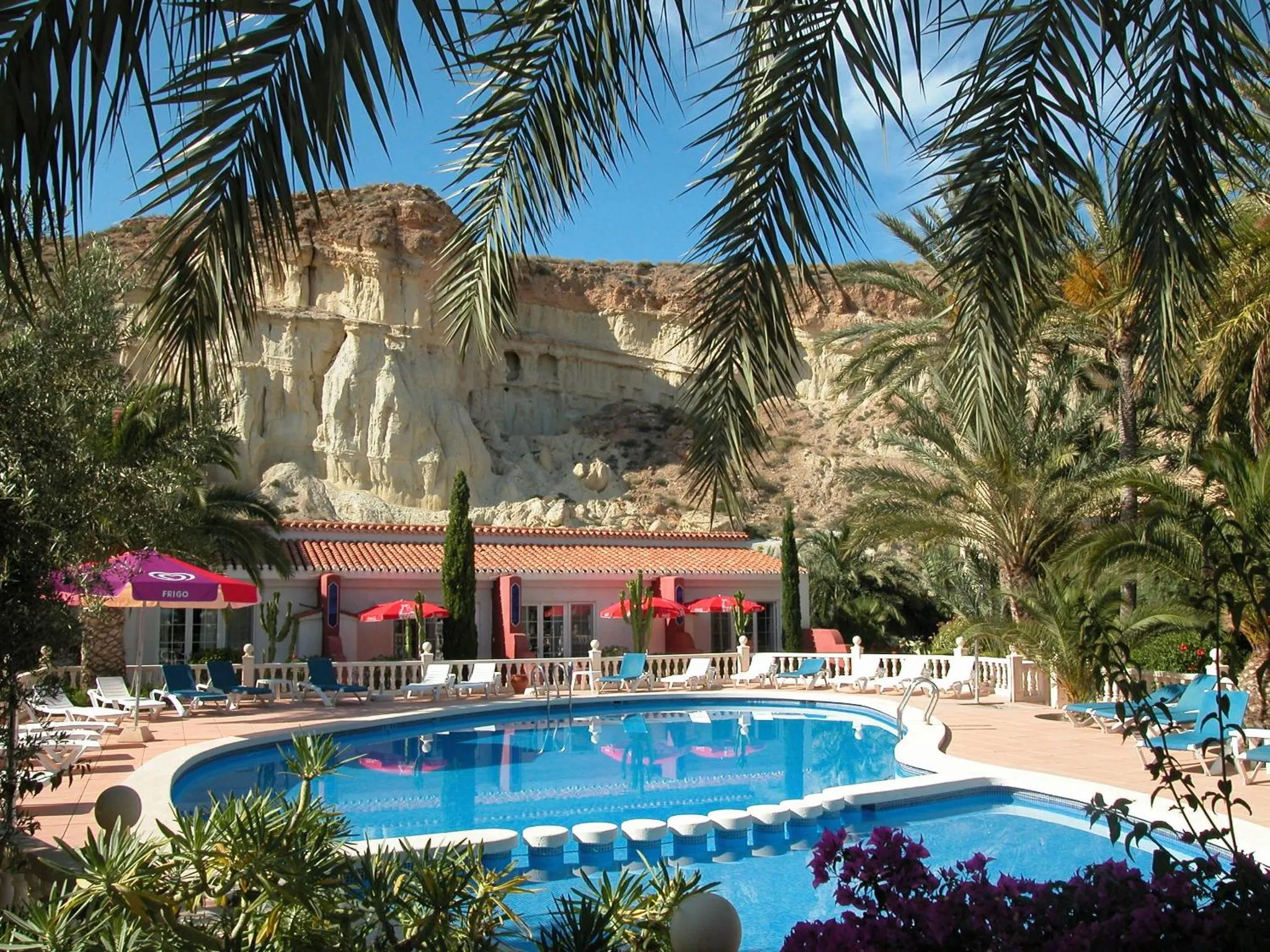Une piscine bordée de transats et de palmiers, avec un bâtiment en arrière-plan et des falaises rocheuses.