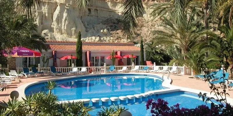 Une piscine rectangulaire bordée de transats et de parasols, devant un bâtiment aux toits en tuiles rouges.