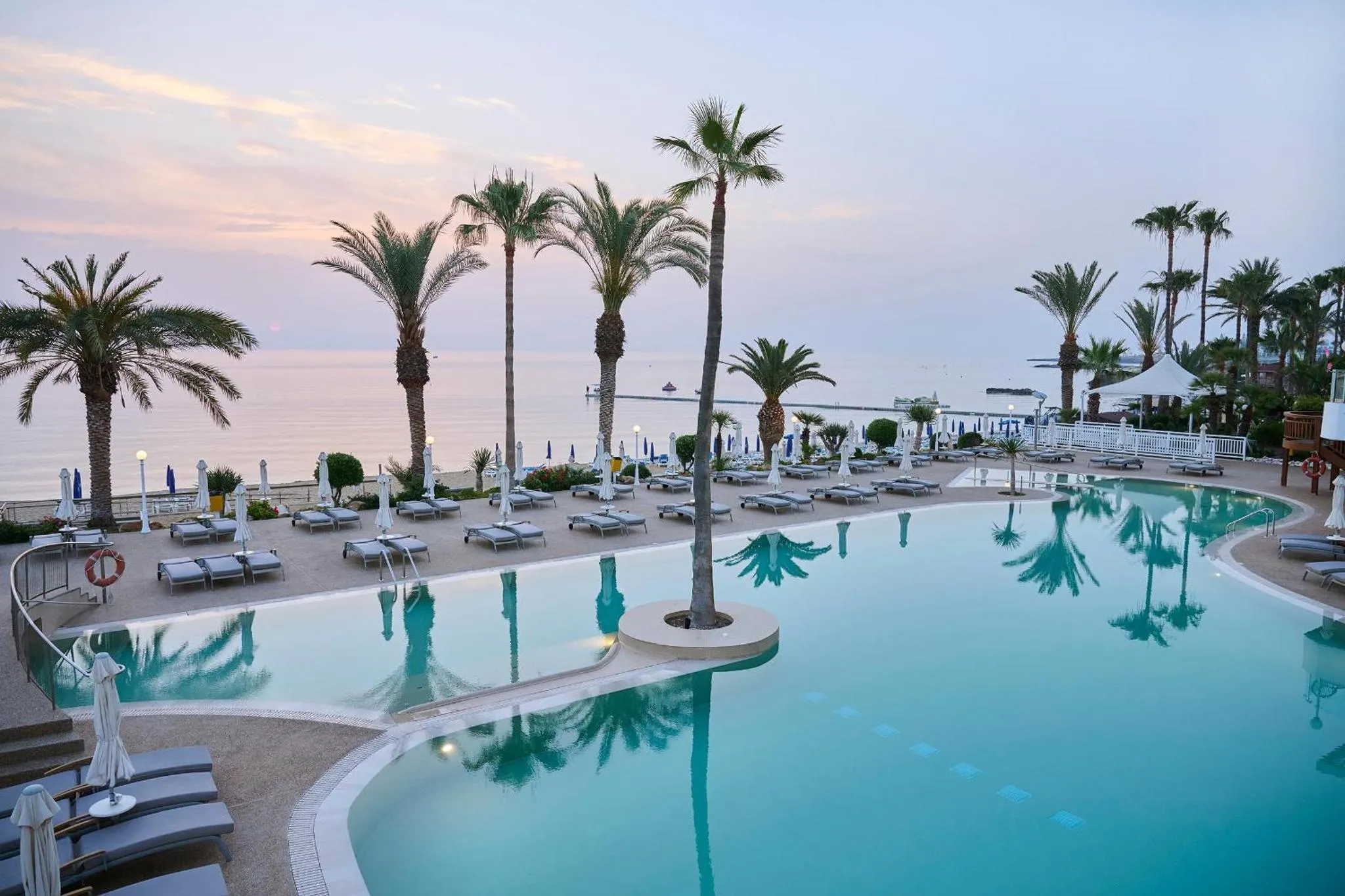 Piscine turquoise bordée de transats, avec palmiers et vue sur la mer au coucher du soleil.