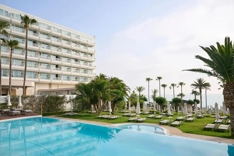 Vue d'un bâtiment blanc avec des balcons, entouré de végétation et d'une piscine.