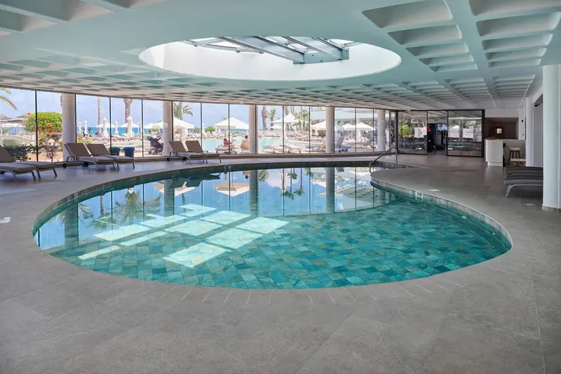 Une piscine intérieure circulaire avec un sol carrelé et une grande baie vitrée donnant sur une plage.