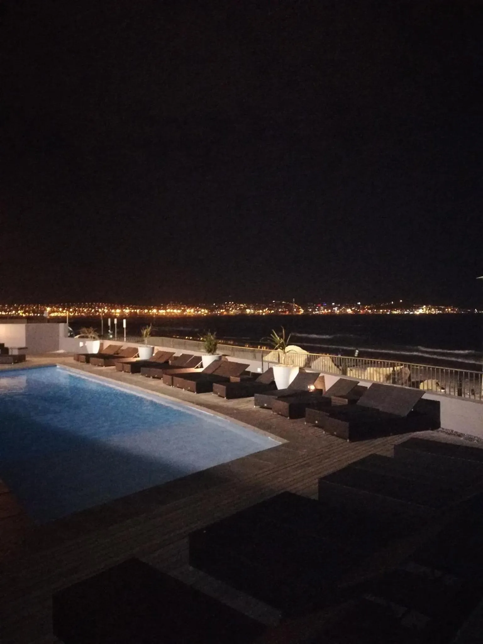 Terrasse lumineuse avec transats et piscine au premier plan, vue sur une ville illuminée en arrière-plan.