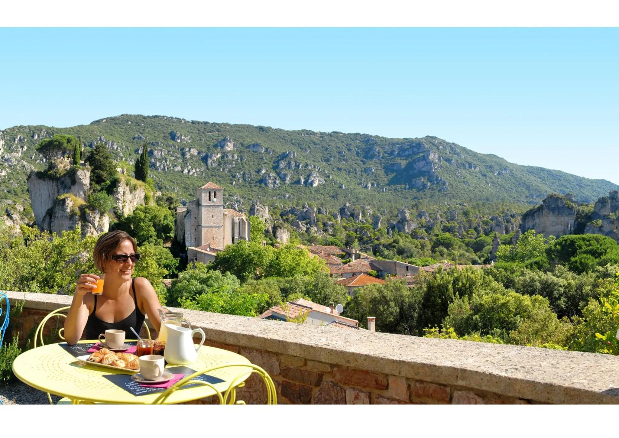 Une personne prend son petit-déjeuner sur une terrasse ensoleillée avec vue sur un paysage vallonné.