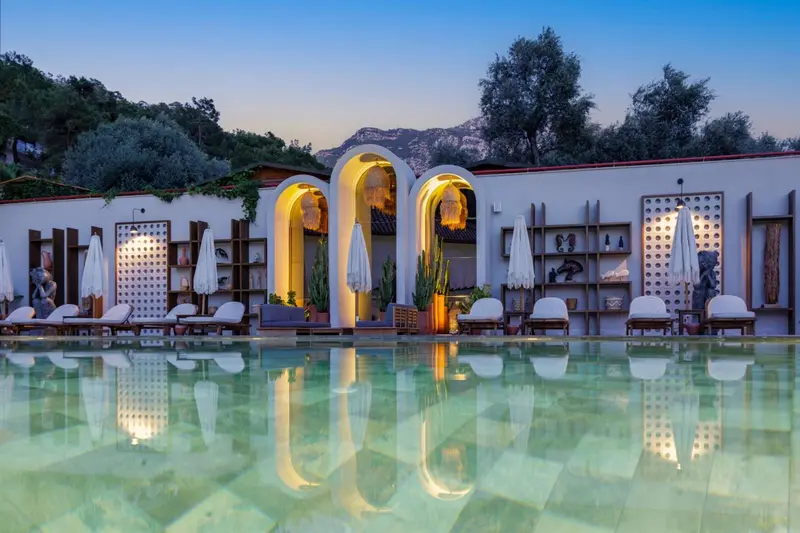 Façade illuminée d'un bâtiment blanc avec des arcades et une piscine au premier plan.