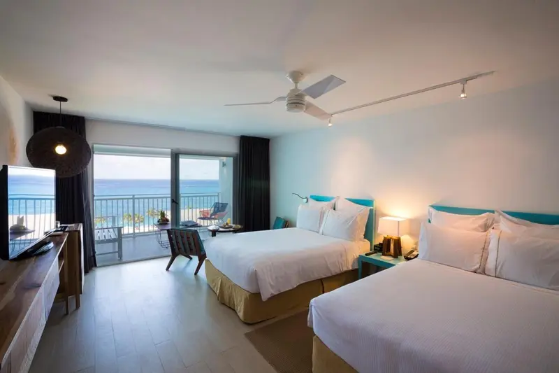 Chambre lumineuse avec deux lits et vue sur la mer depuis un balcon.