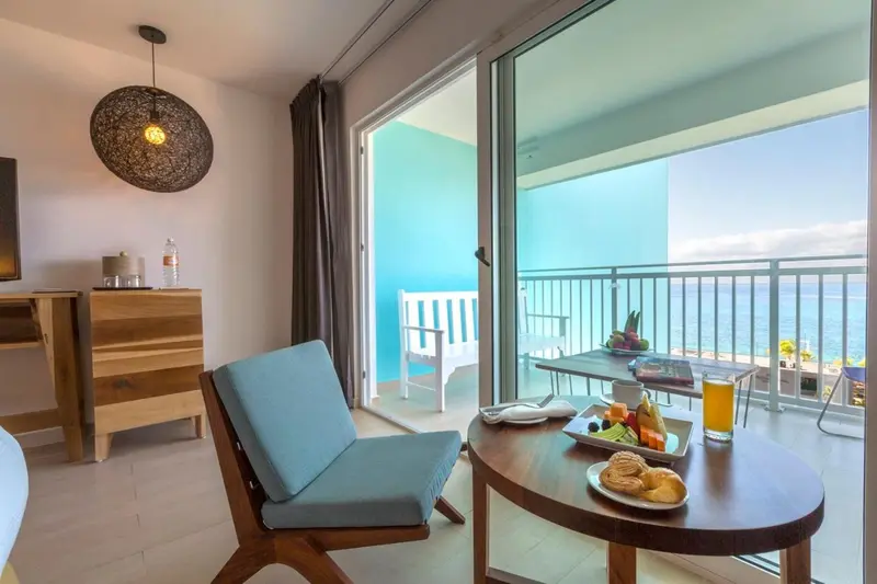 Une chaise et une table basse avec des fruits et une boisson sont disposées devant une baie vitrée donnant sur une terrasse.
