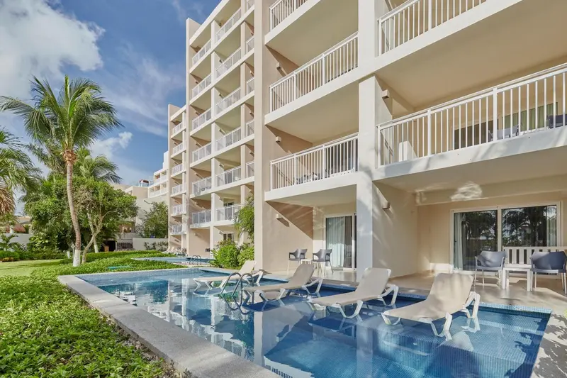 Une piscine bordée de chaises longues devant un bâtiment beige à balcons.