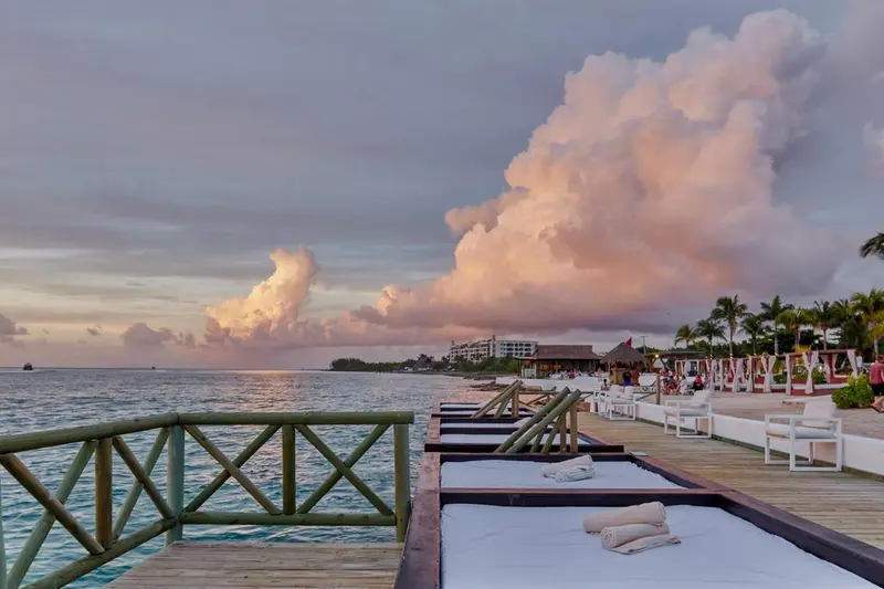 Un ponton en bois mène vers des transats blancs sur une plage au coucher du soleil.
