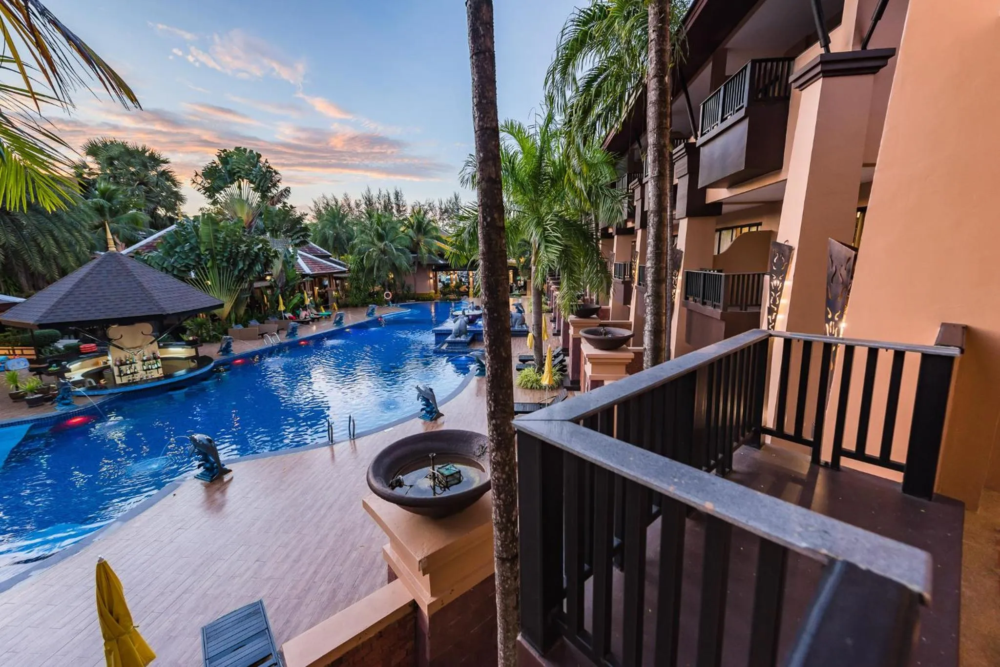Vue d'une piscine d'hôtel avec des transats et des palmiers au coucher du soleil.
