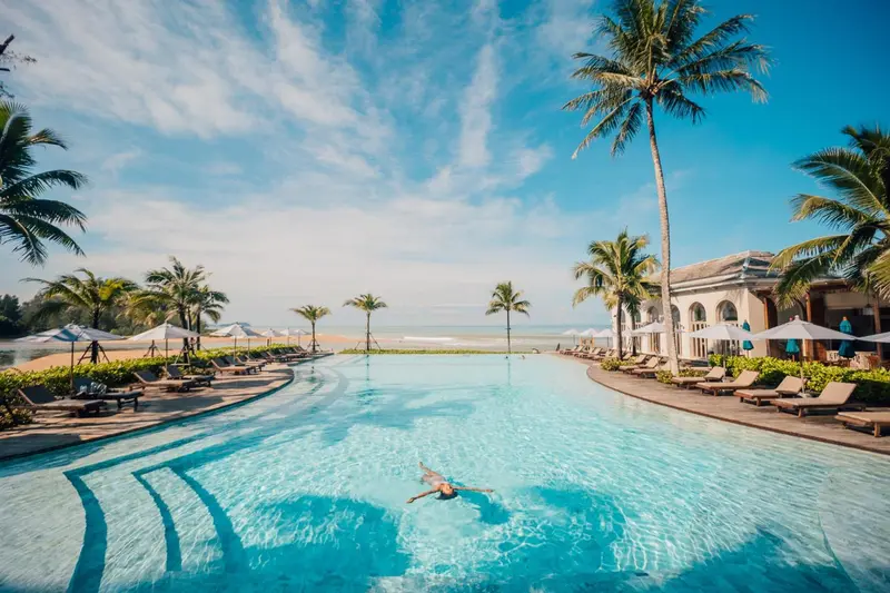Une piscine turquoise bordée de transats, avec une plage en arrière-plan et des palmiers.