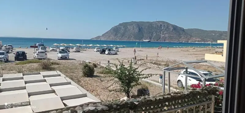 Vue sur une plage de sable fin et une mer bleue, avec des parasols et des véhicules à l'arrière-plan.