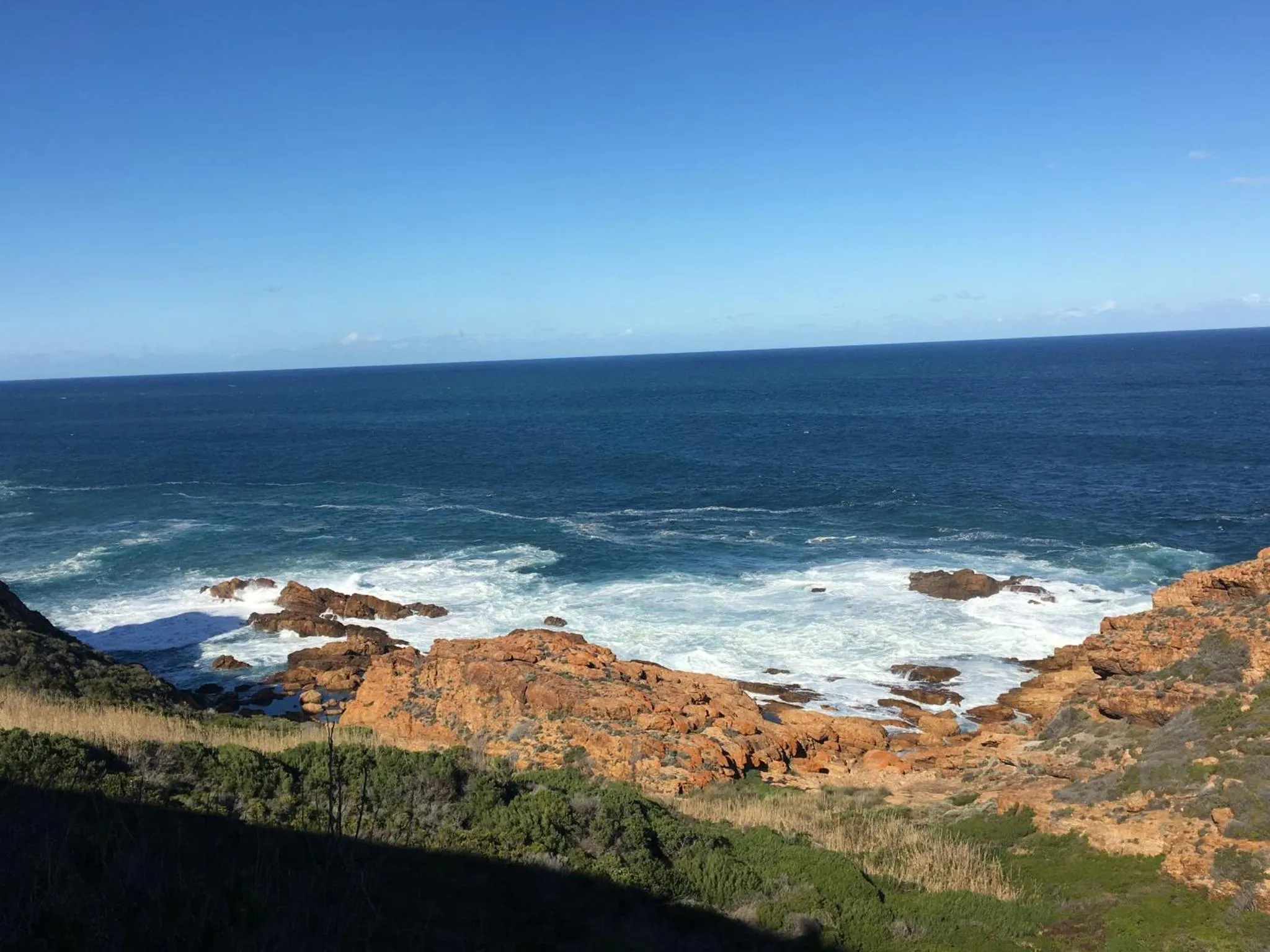 Vue côtière avec des vagues déferlant sur des rochers rouges et une mer bleue.