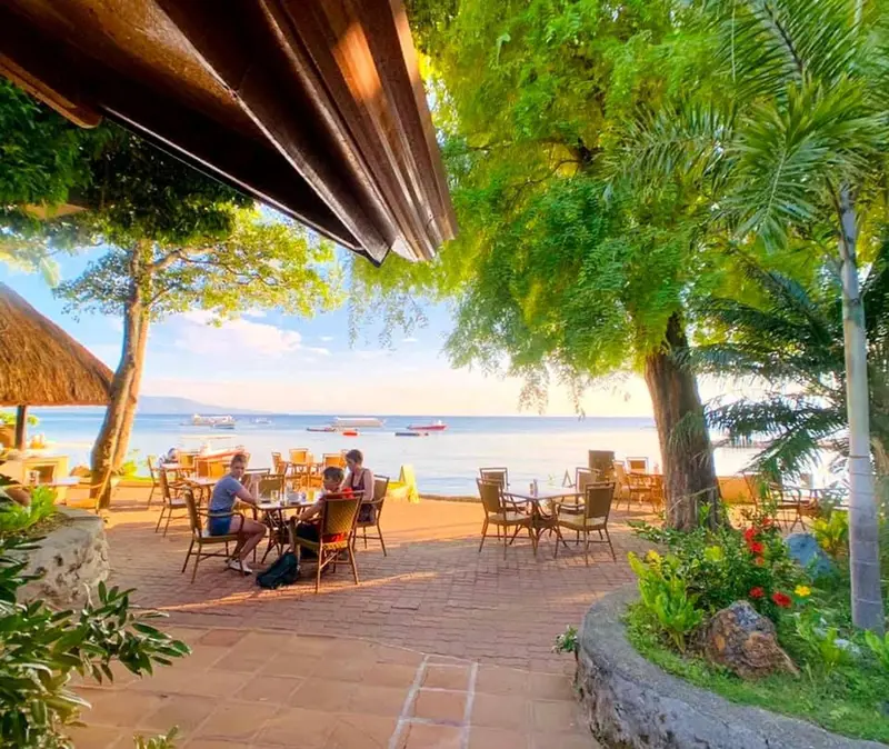 Terrasse ombragée avec tables et chaises donnant sur la mer au coucher du soleil.