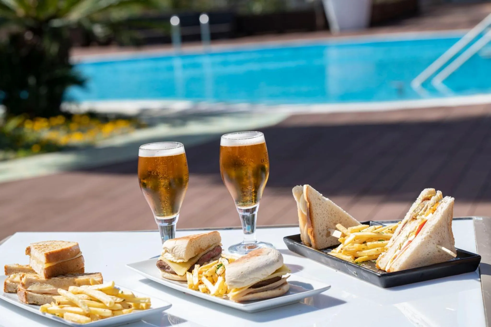 Deux verres de bière et une assiette de sandwichs et de frites sur une table près d'une piscine.