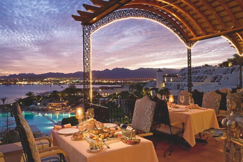 Tables dressées en terrasse avec vue sur la mer et les montagnes au coucher du soleil.
