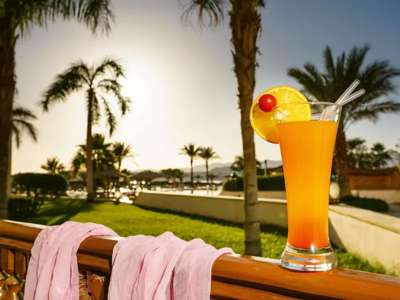 Un cocktail coloré est présenté sur une rambarde en bois avec une serviette rose, devant une végétation luxuriante et un ciel clair.