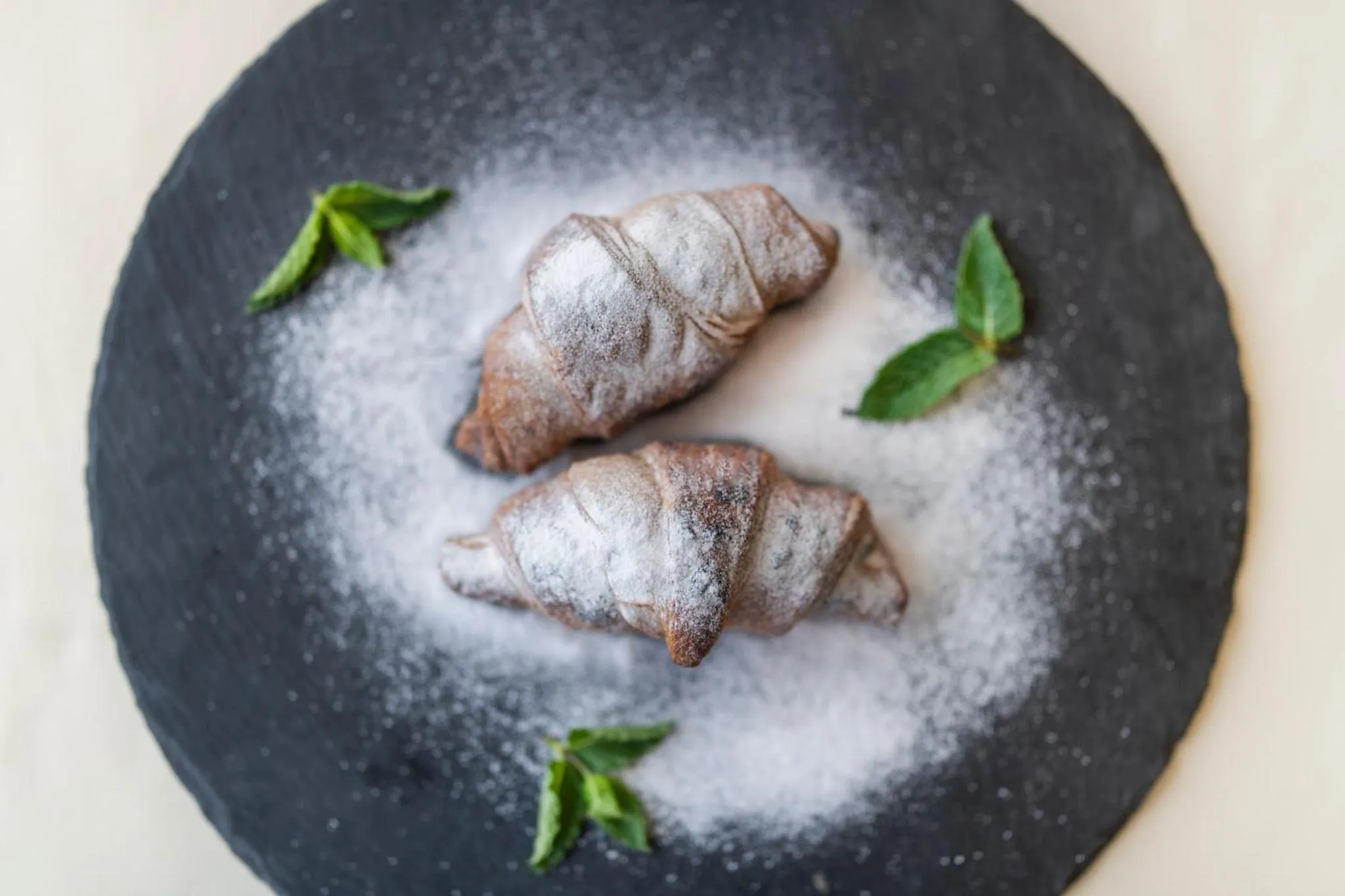 Deux croissants au chocolat saupoudrés de sucre glace sont présentés sur une ardoise noire avec quelques feuilles de menthe.