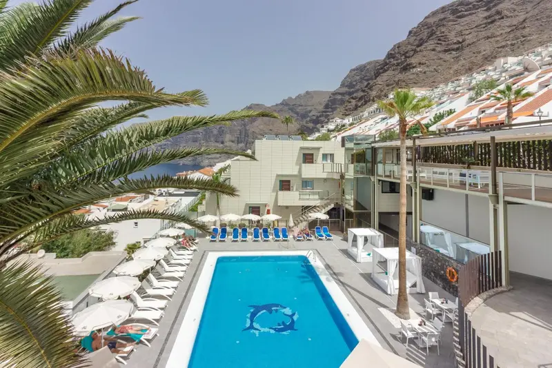 Piscine rectangulaire entourée de chaises longues et de parasols, avec un bâtiment blanc en arrière-plan et des montagnes au loin.