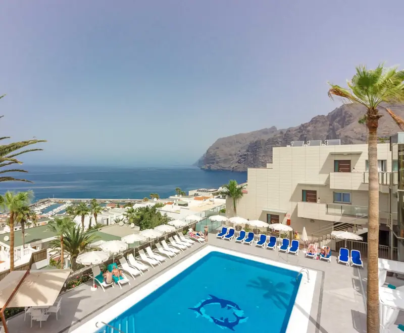 Vue aérienne d'une piscine d'hôtel avec transats et palmiers, face à la mer et aux montagnes.