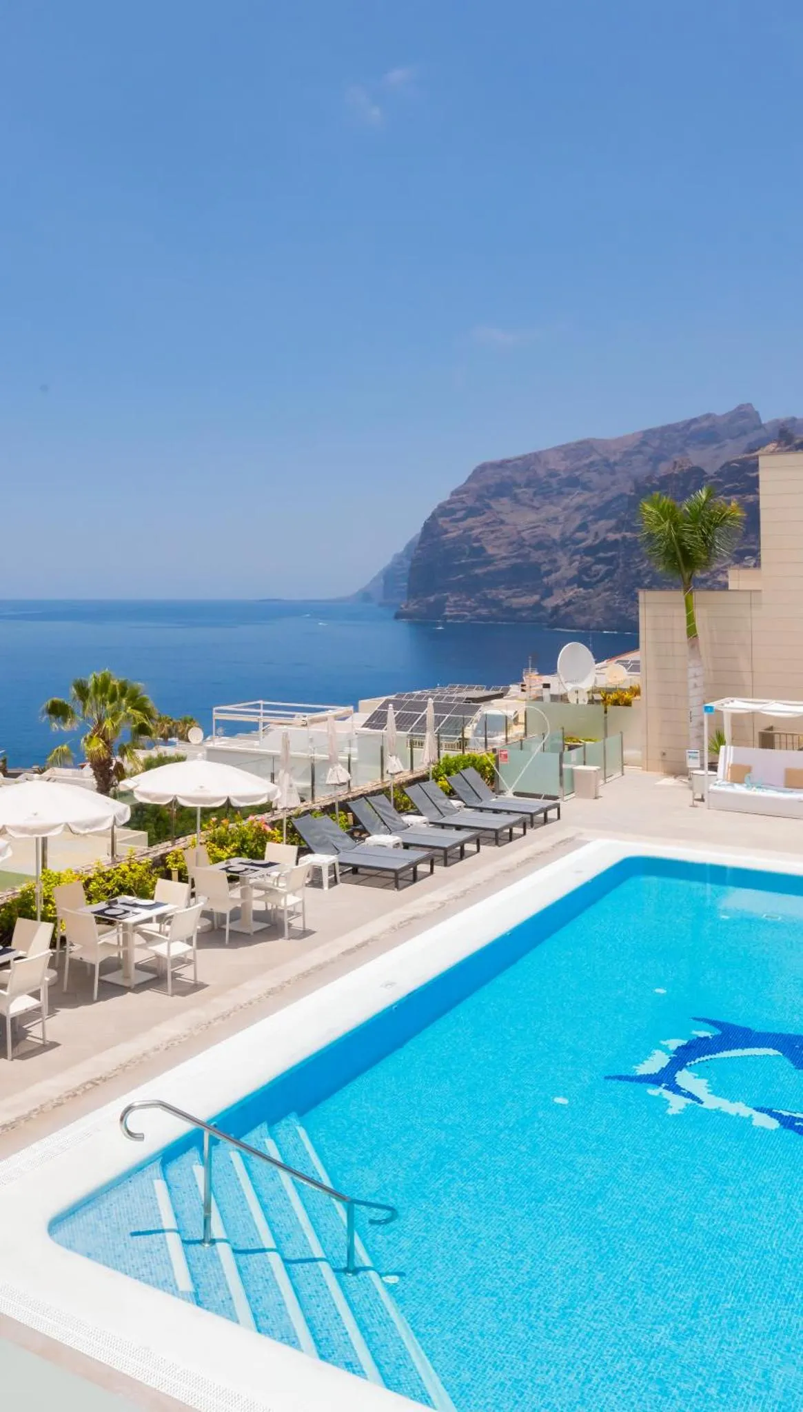 Une piscine turquoise bordée de transats blancs offre une vue sur la mer et les collines.